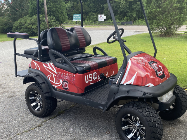 Red off-road golf cart with oversized tires parked on grass near a road