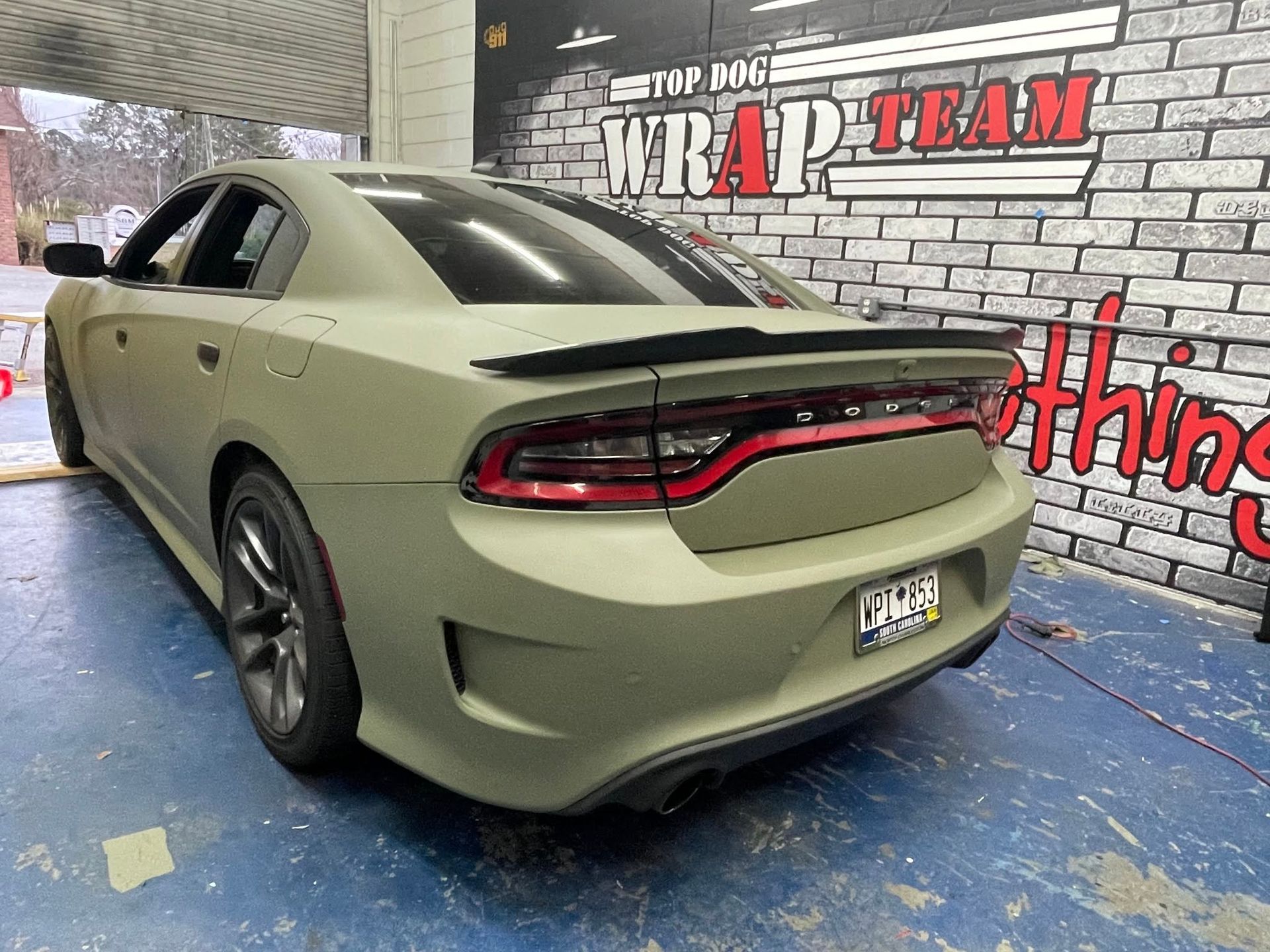 Olive green Dodge Charger in a garage, rear three-quarter view, with black spoiler and taillight bar.