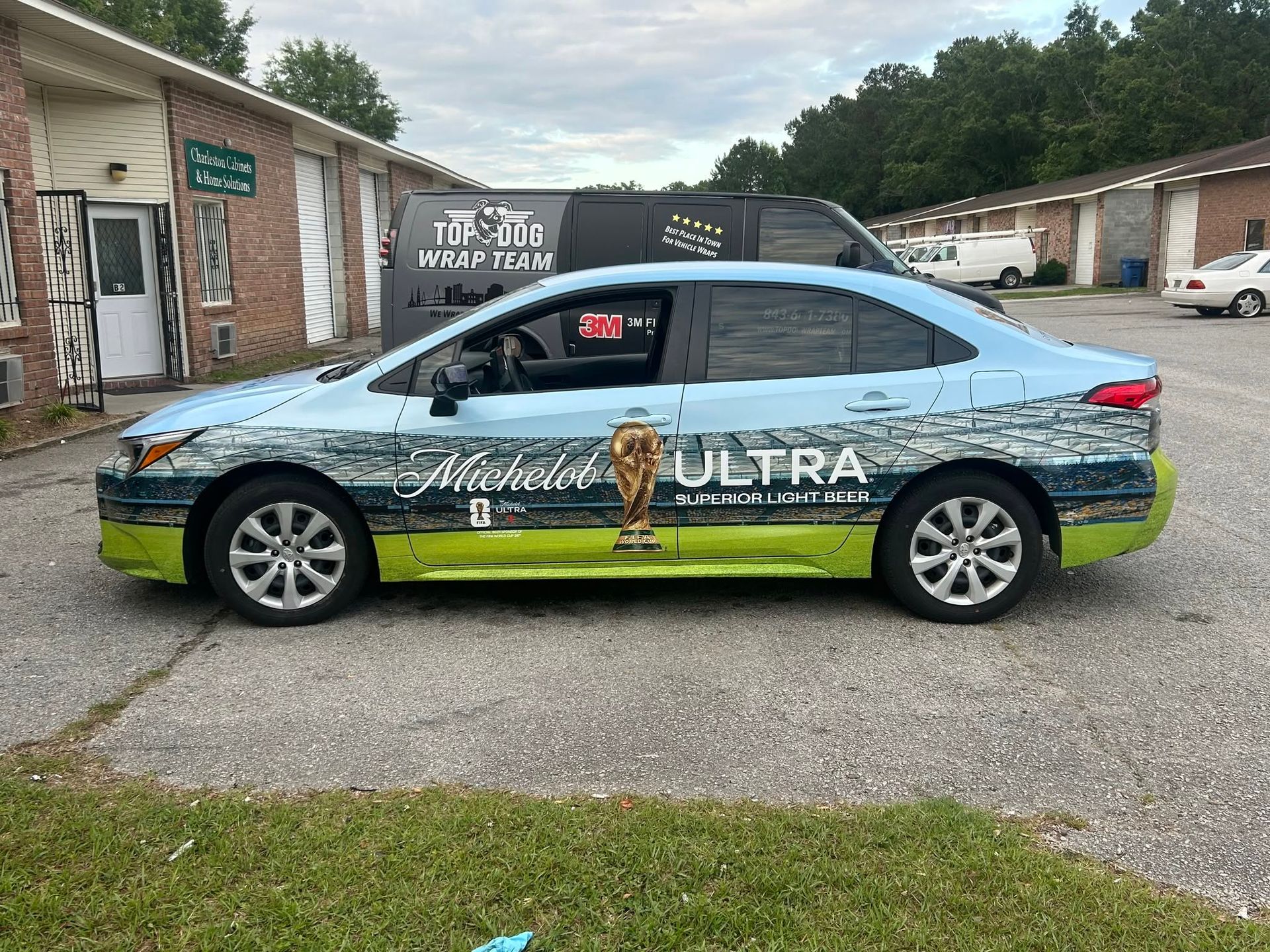 Wrapped blue-and-green car parked in a lot beside brick buildings