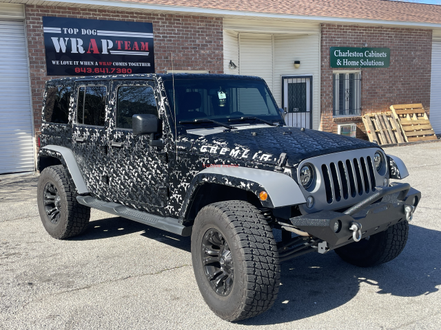 Black Jeep Wrangler parked outside a wrap shop, with black camo wrap and off-road tires
