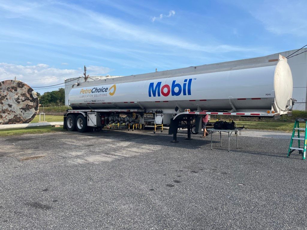 Mobil fuel tanker parked on gravel beside a road with two workers nearby under a blue sky