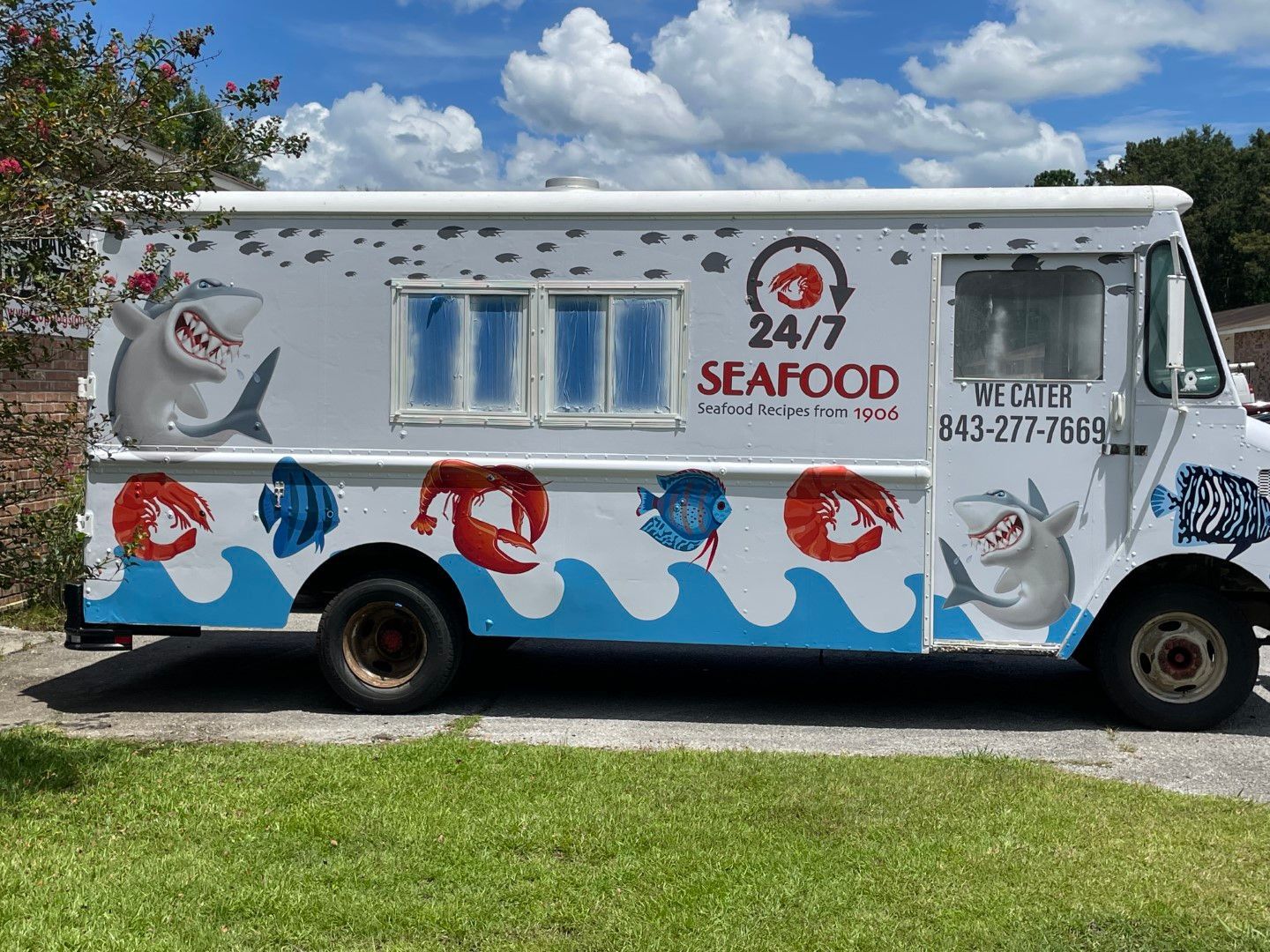24/7 seafood truck with blue wave and fish mural parked on grass under a cloudy sky