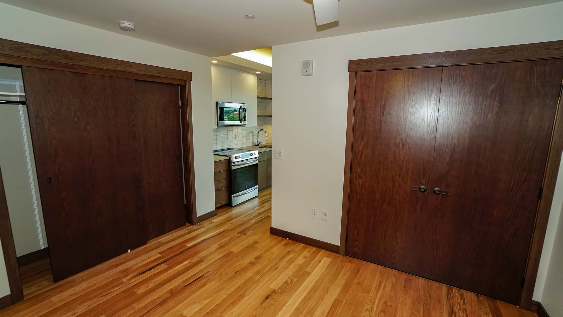 An empty room with hardwood floors and sliding doors leading to a kitchen.