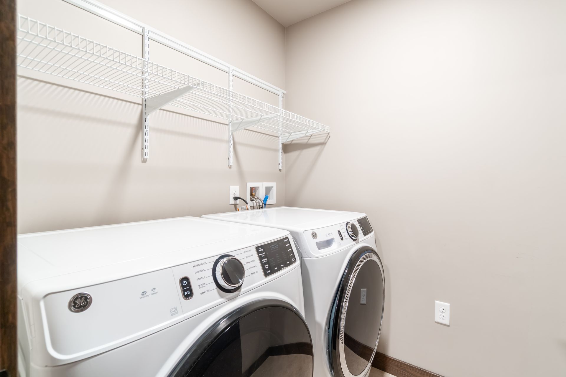 Laundry room with white washer and dryer, wire shelves, and beige walls.