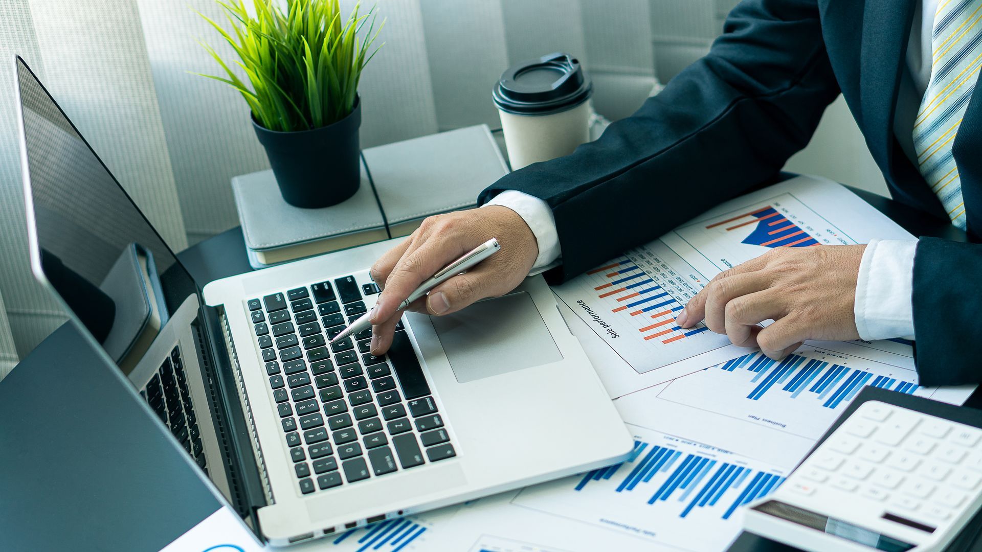 Person working at a laptop with printed charts and graphs on a desk, in an office setting