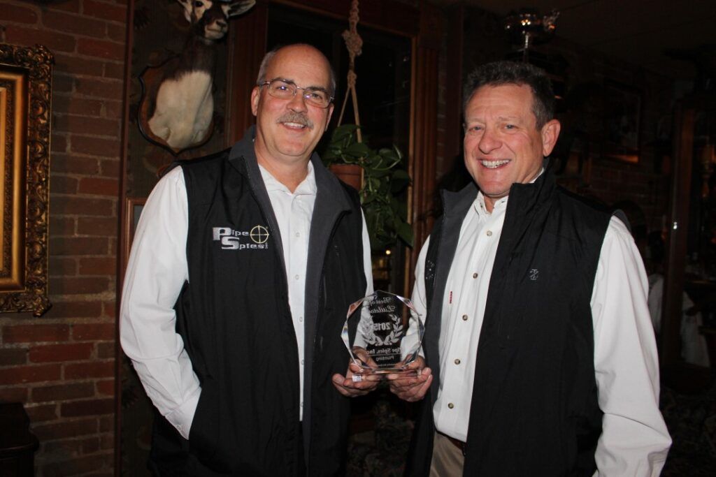 Two men in vests pose, one holding an award. They stand inside a room with brick walls and artwork.