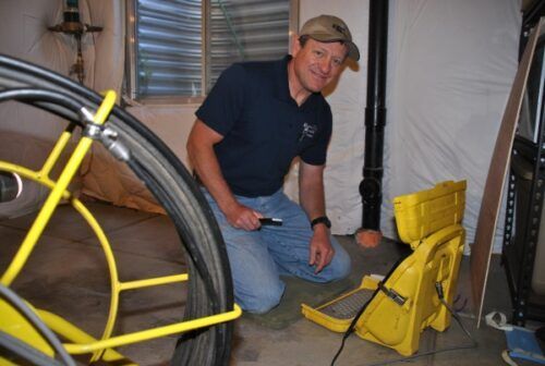 Man kneels in basement holding tool, near yellow equipment and coiled cable.