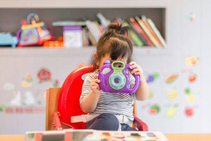 A little girl is sitting in a high chair holding a toy camera at Thornlie Early Learning Centre.