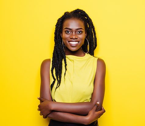 A woman with dreadlocks is smiling with her arms crossed against a yellow background.