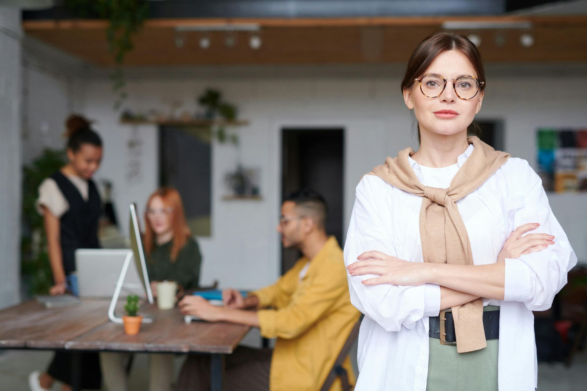 Woman standing with her arms folded looking at the camera, her colleagues are chatting in the background.