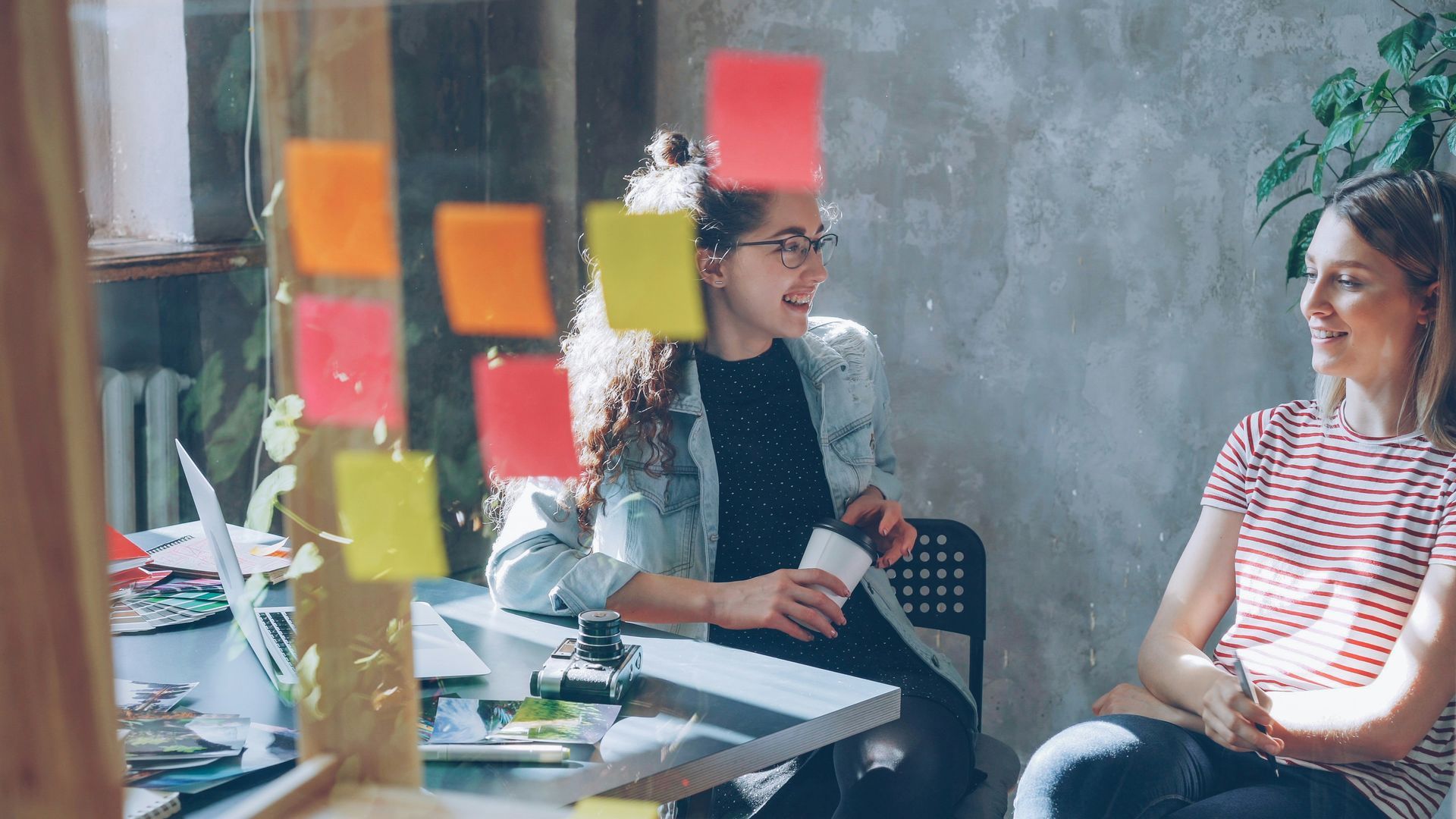 Two people talking in a bright office, with colorful sticky notes on a glass wall.