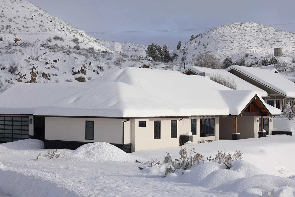 House covered in snow, with snowy mountains in the background. The house is white and beige with a glass garage.