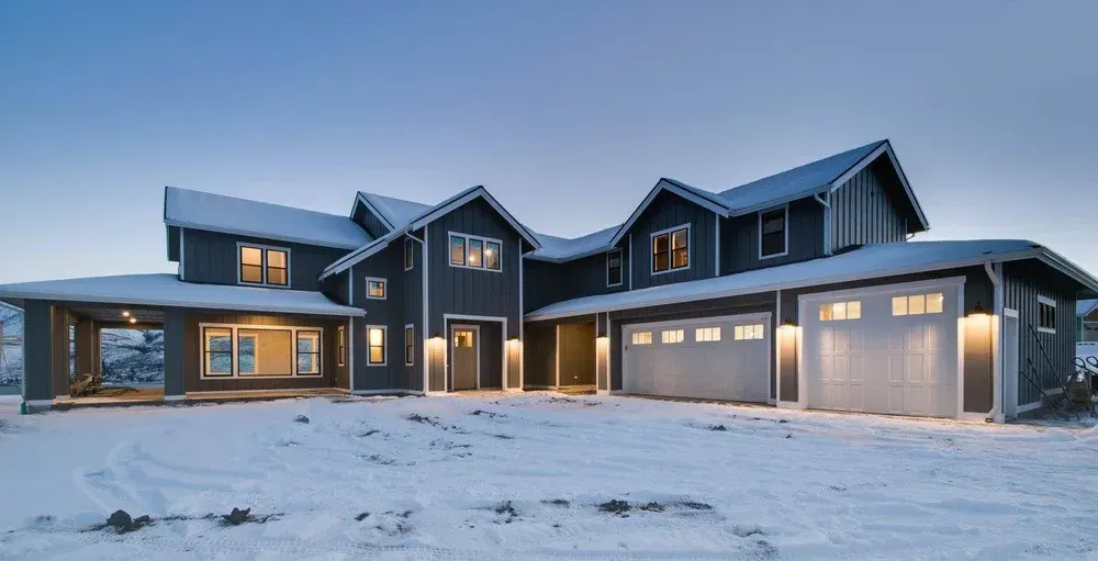 Two-story gray house with white garage doors and a snowy yard at dusk.