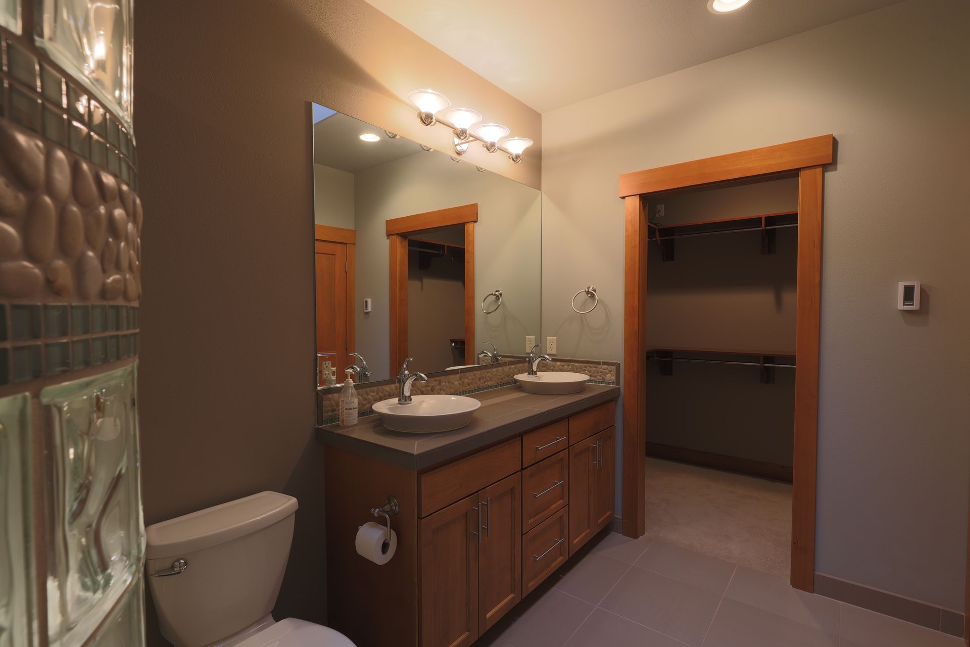 Bathroom with double sinks, wooden cabinets, toilet, and walk-in closet with shelves.