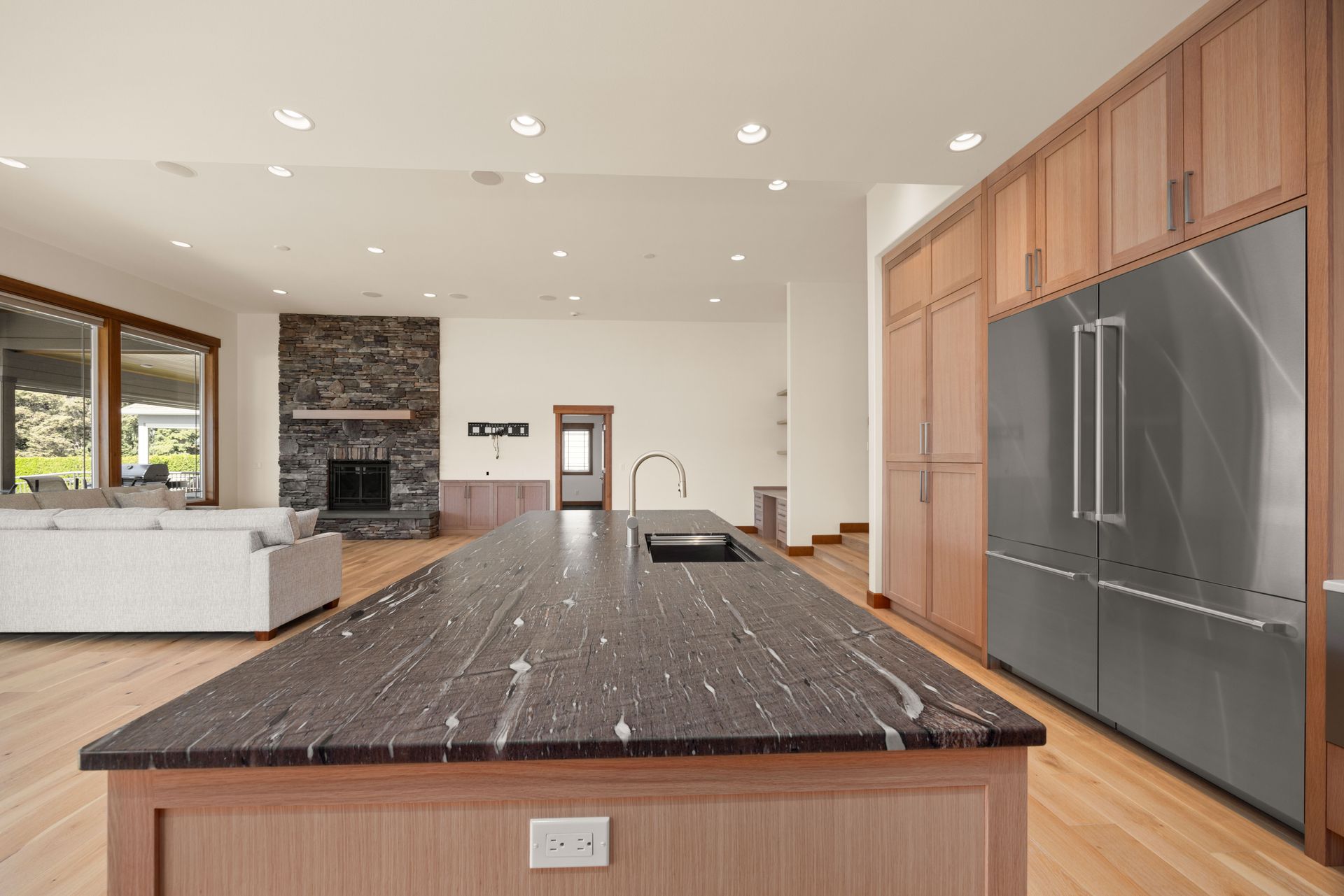 Modern kitchen with large island, stainless steel refrigerator, and stone fireplace in background.
