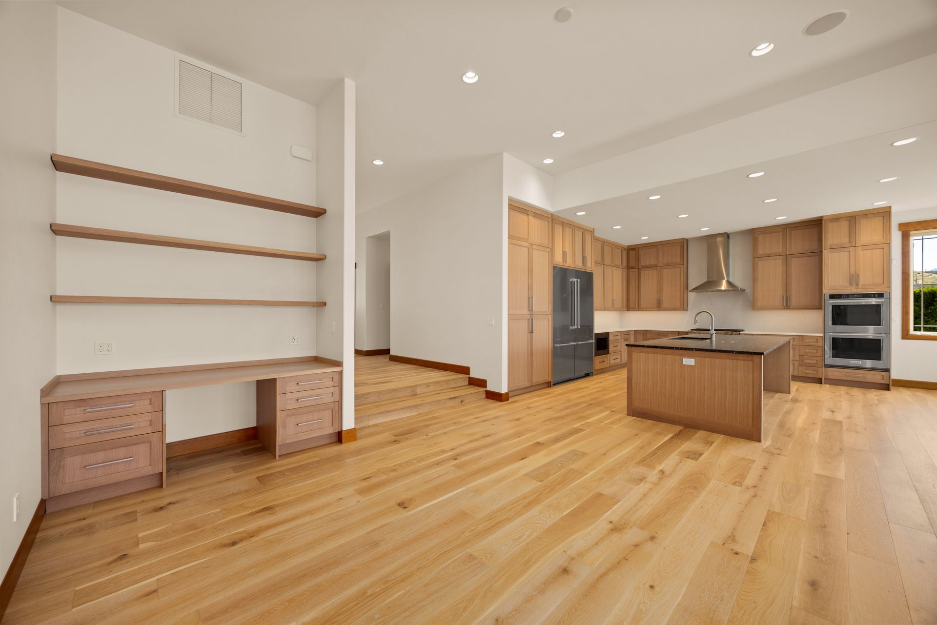 Open-plan kitchen and desk area. Wooden cabinets, island, and flooring. Stainless steel appliances. White walls.