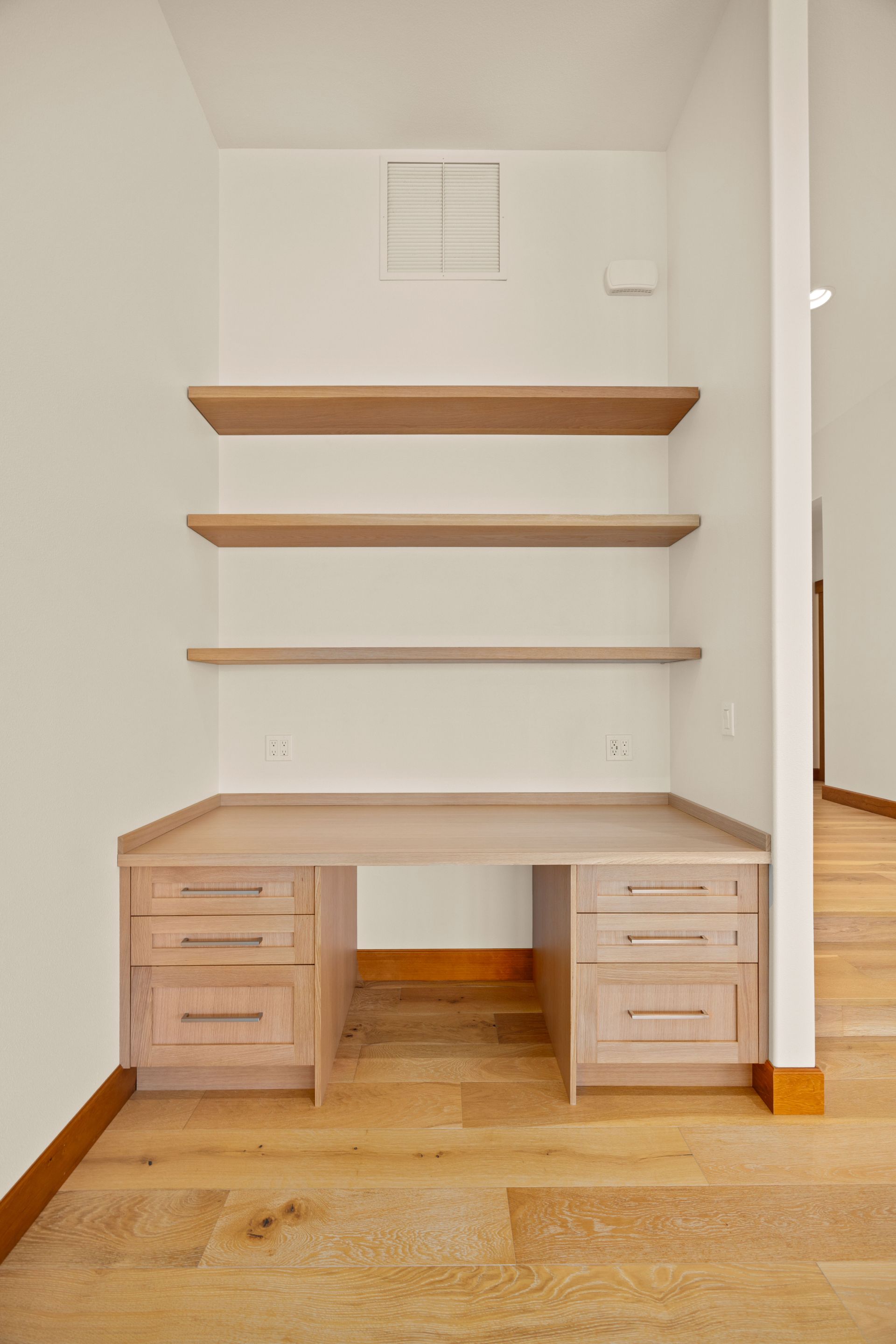 Built-in desk with three shelves, light wood finish, against a white wall in a room with wood flooring.