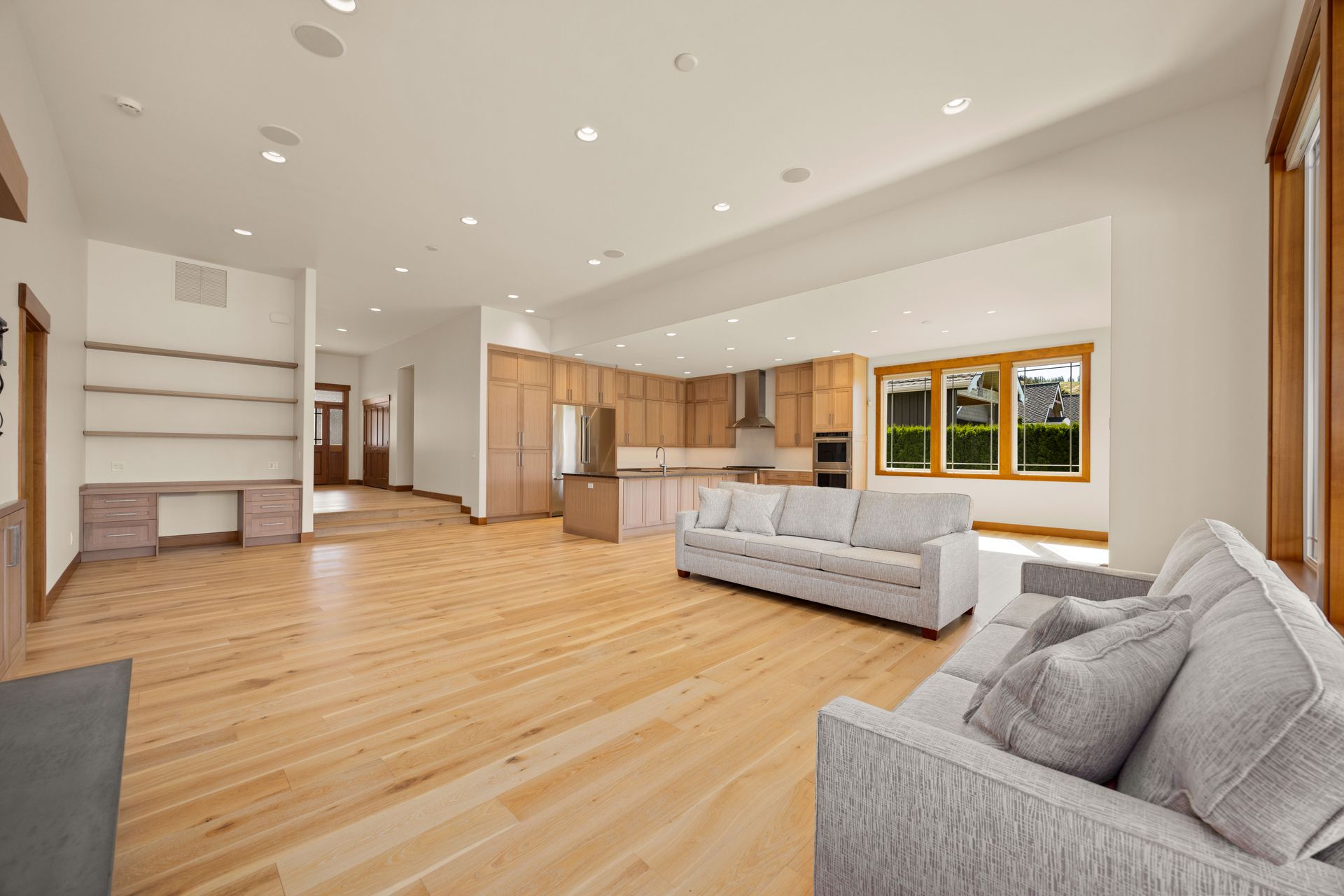 Spacious, open-plan living area with blonde wood floors, light gray sofas, and kitchen with wood cabinets.