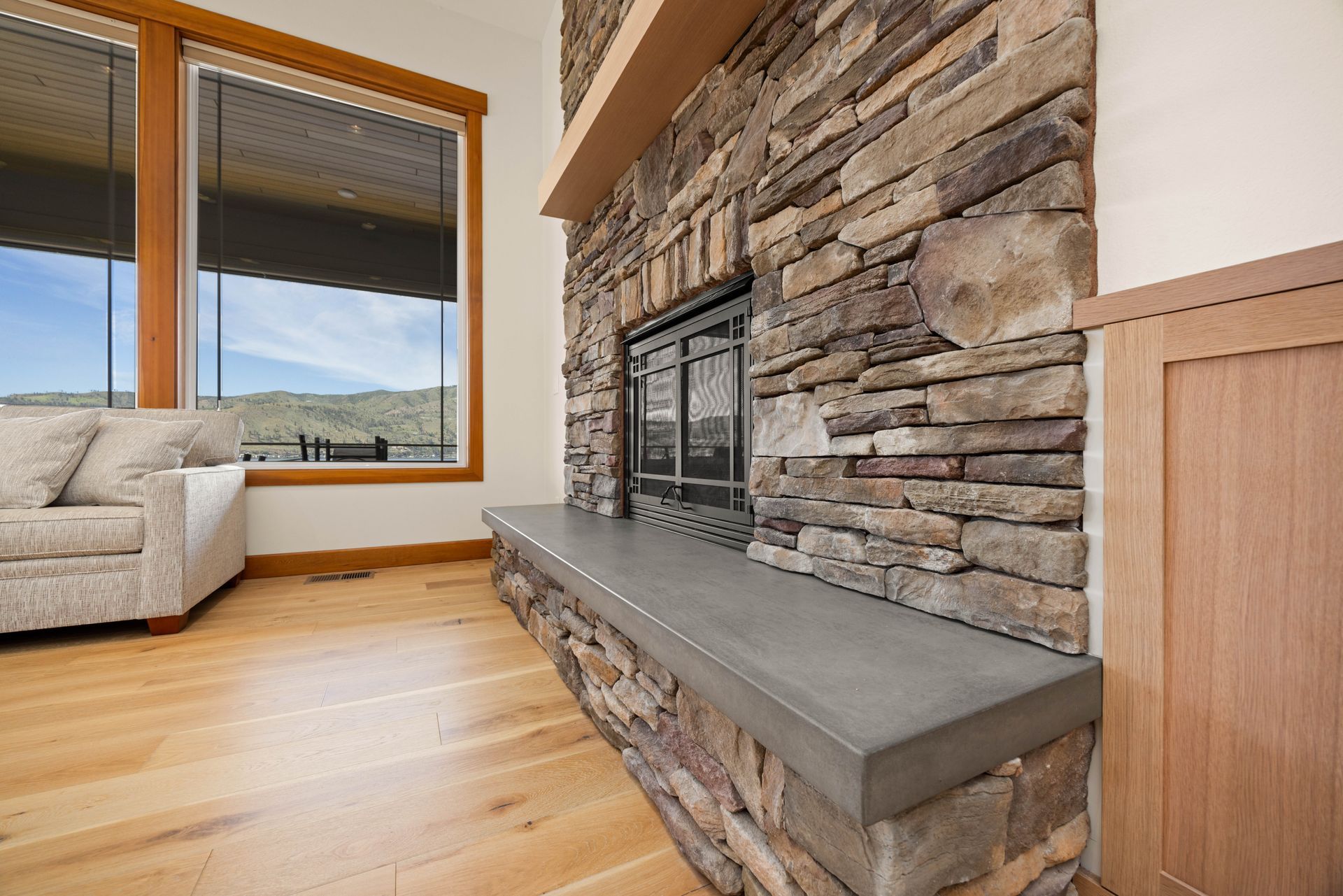 Fireplace with stone facade, dark mantel, concrete hearth, wooden floor and adjacent window.