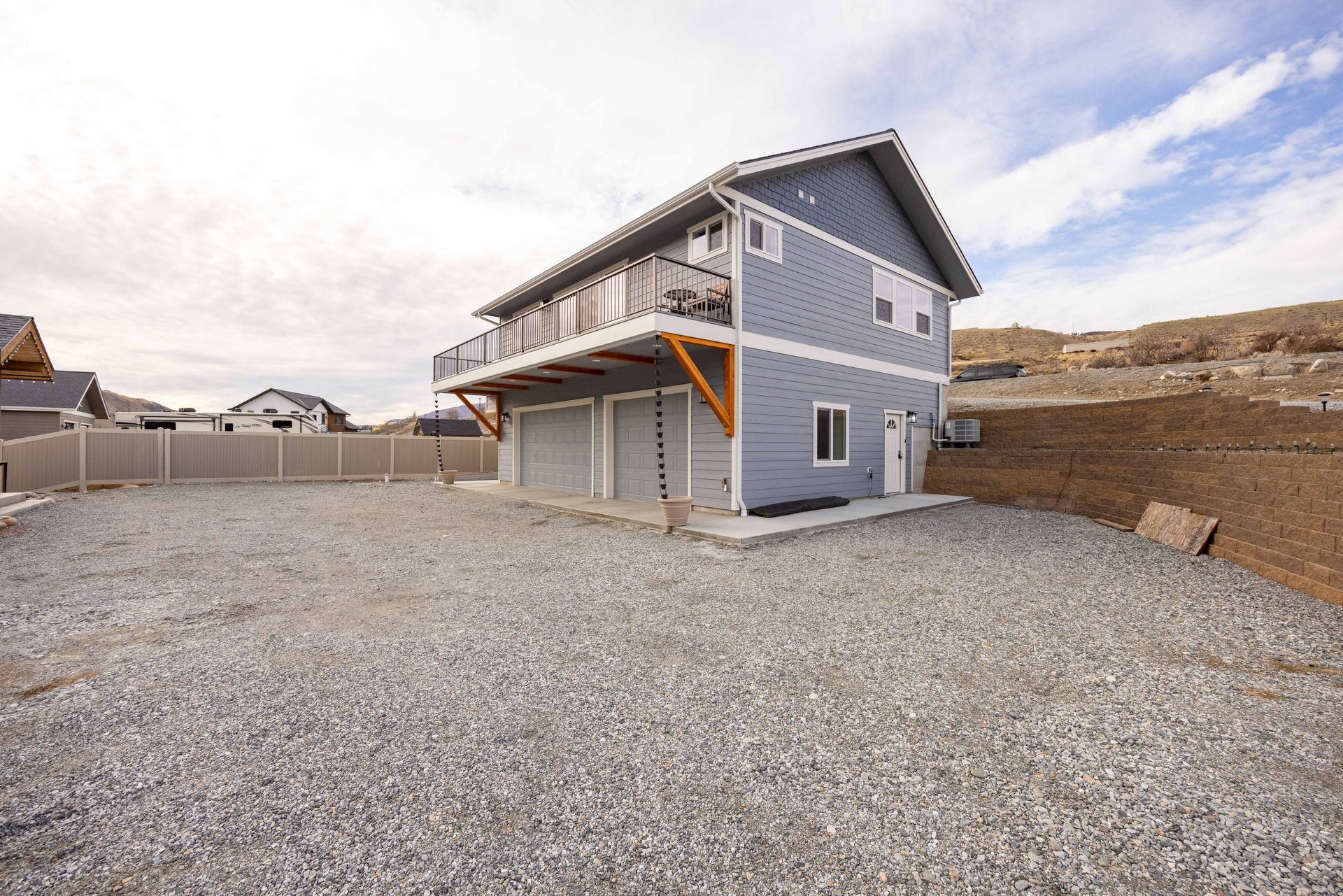 Blue two-story house with a balcony over a three-car garage on a gravel lot.