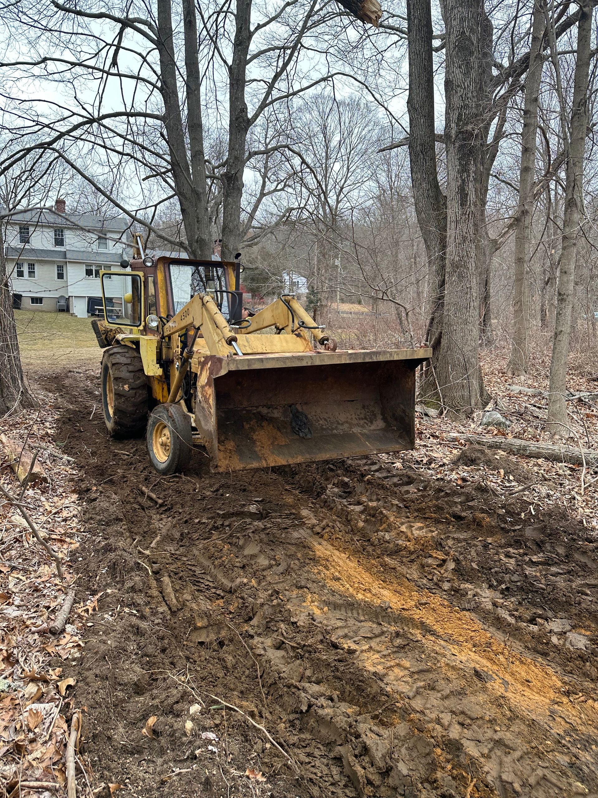 Yellow backhoe clearing a muddy wooded path beside a house in winter