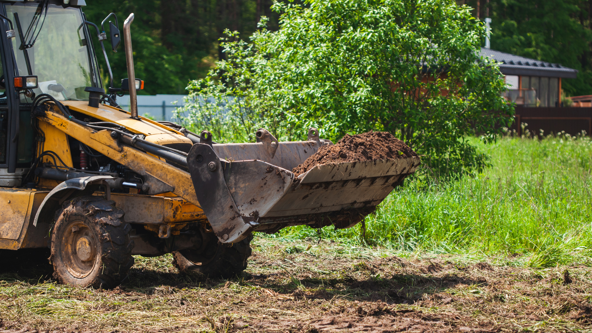 Yellow backhoe loader with raised bucket on muddy ground near green trees and a building