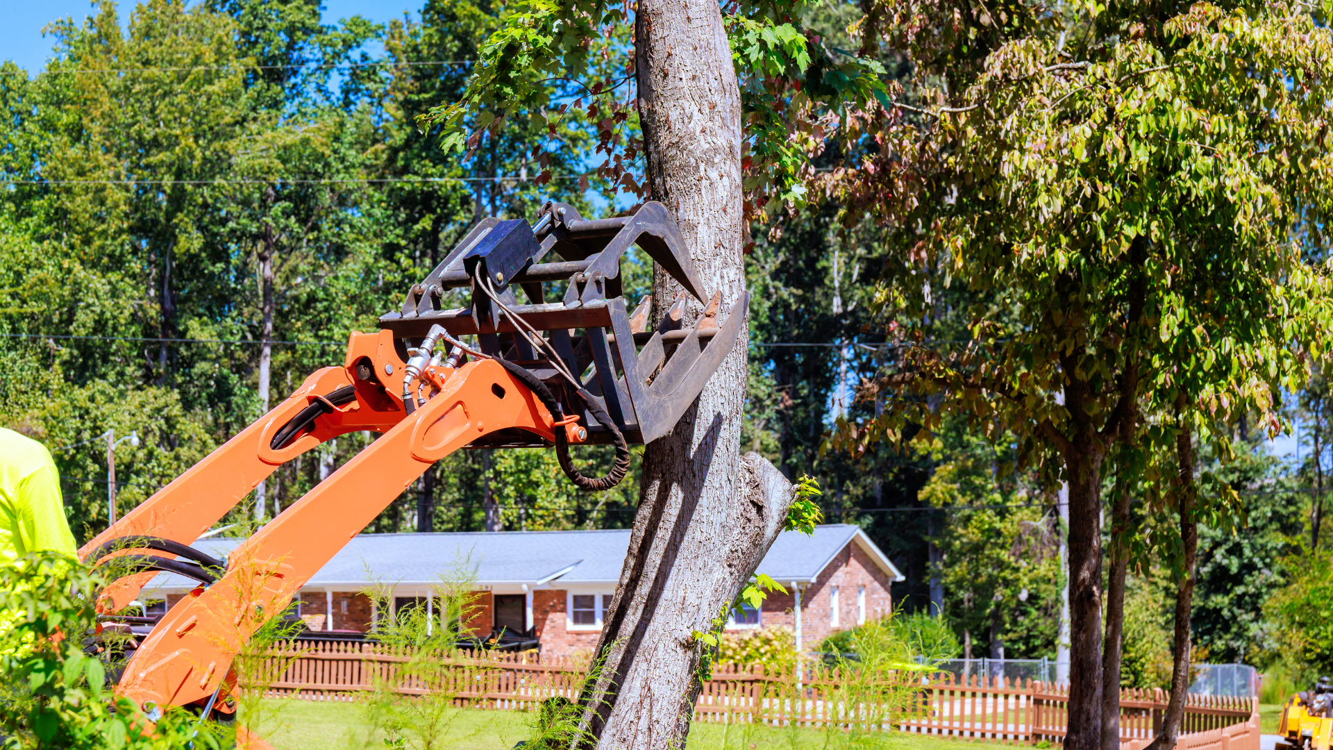 Orange excavator lifting a tree trunk beside a house in a suburban yard