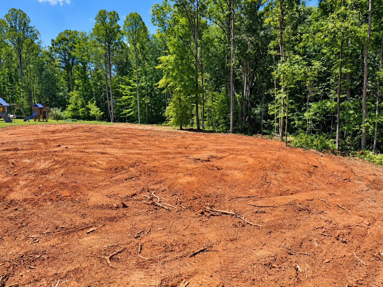 Freshly cleared dirt lot bordered by tall green trees under a bright blue sky