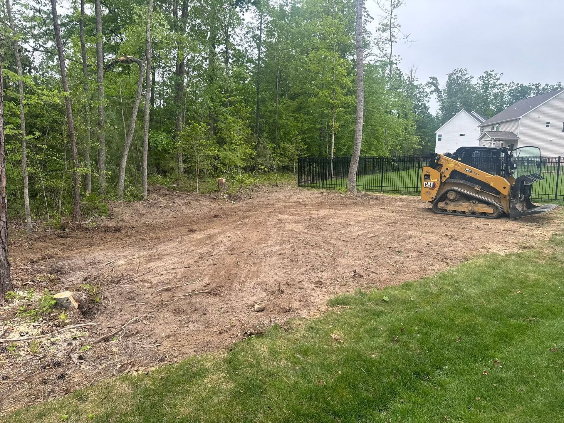 Small excavator grading a dirt clearing beside a grassy yard and trees near a house