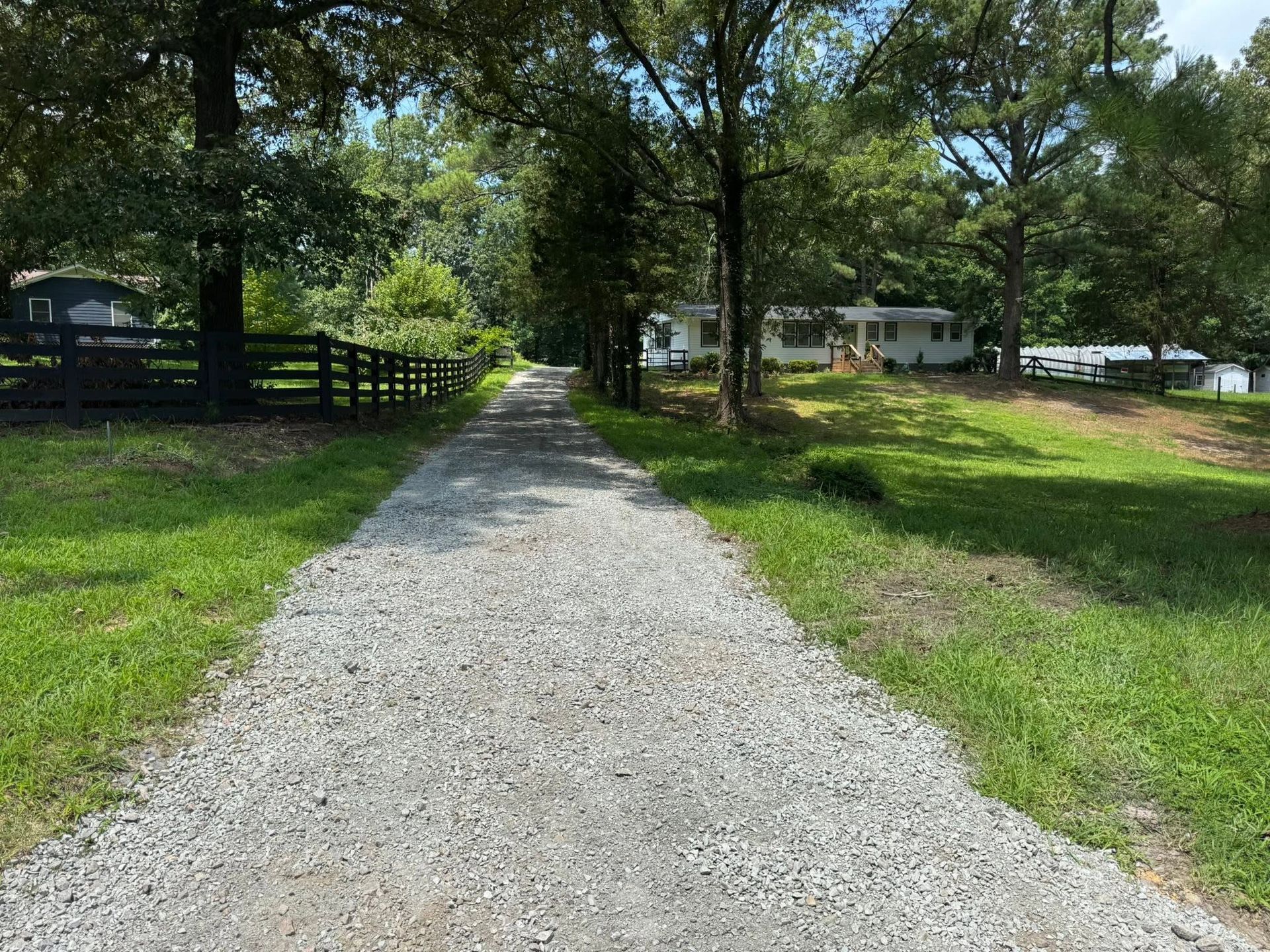 Gravel driveway leading to a white house through a green, tree-lined yard on a sunny day
