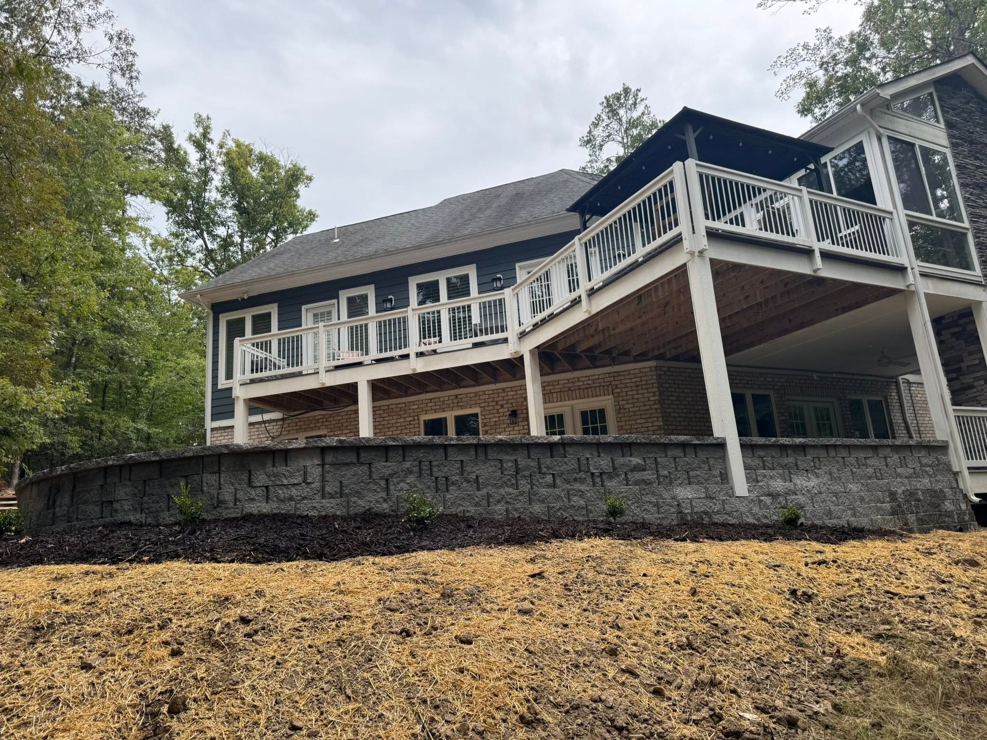 House with stone base and white-railed deck on a wooded hillside, viewed from below