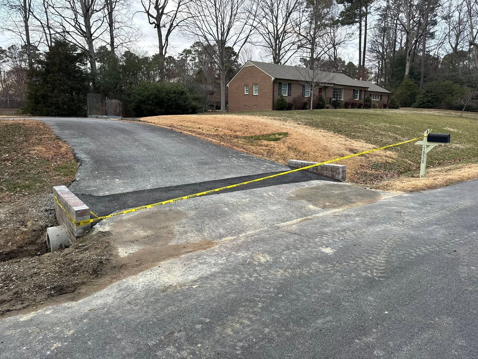 Blocked driveway entrance with a raised curb, yellow caution tape, and a mailbox beside a house on a cloudy day