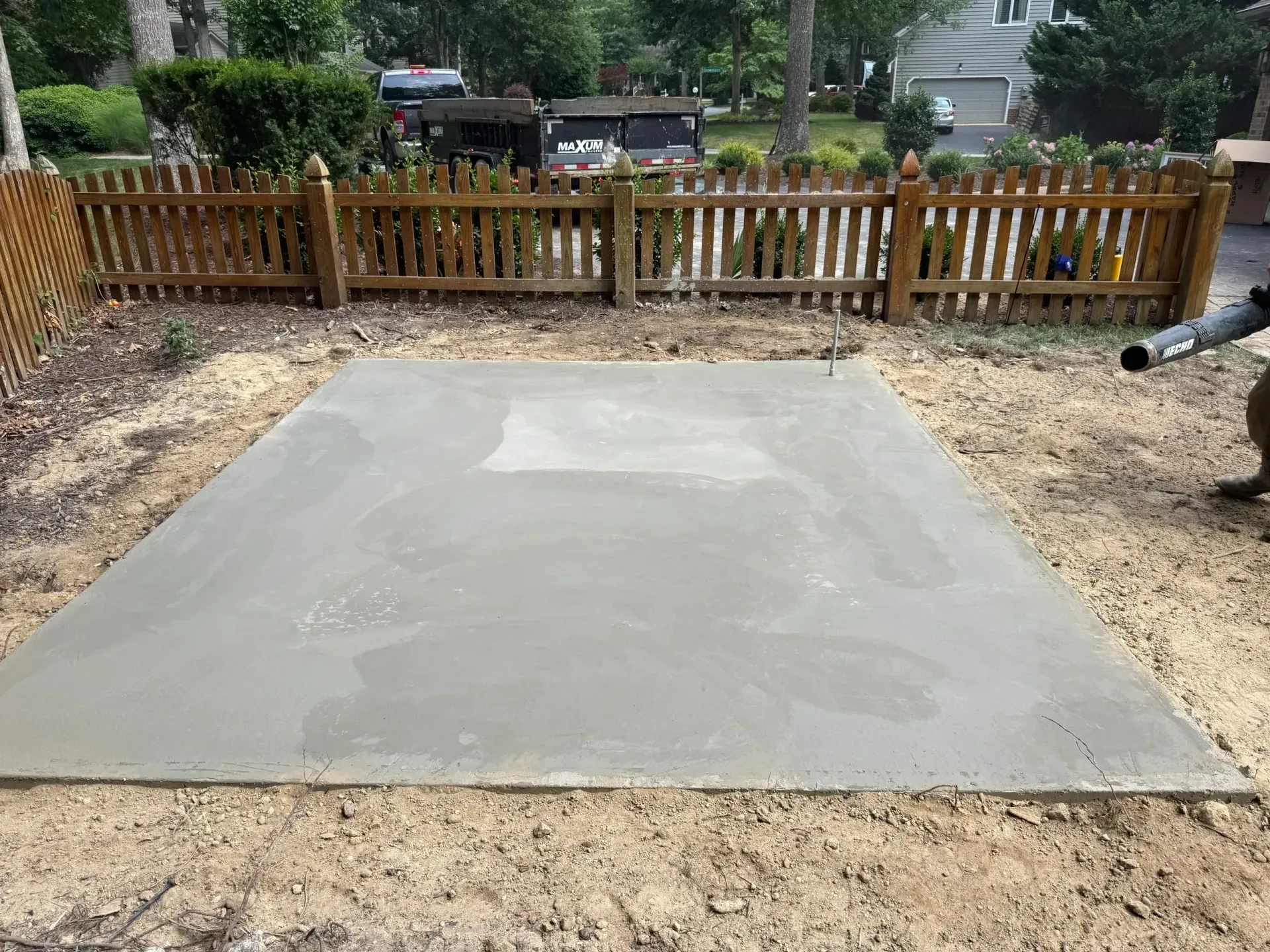 Concrete patio slab in a fenced backyard, surrounded by gravel and trees.