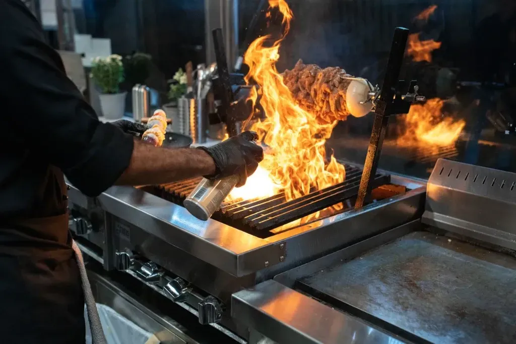 Chef tending a flaming grill in a commercial kitchen, with flames rising from the cooking surface