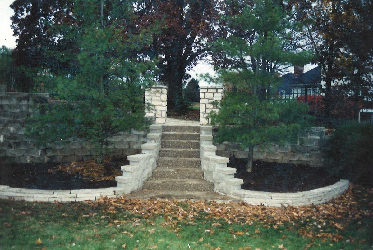 Stone steps lead to an opening between two stone walls, flanked by evergreen trees.