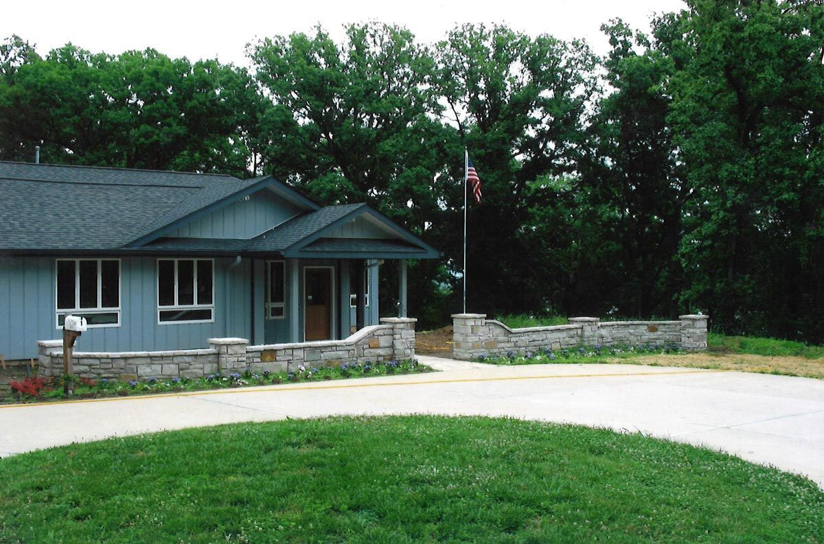Blue building with stone wall and a flag in front of green grass and trees.