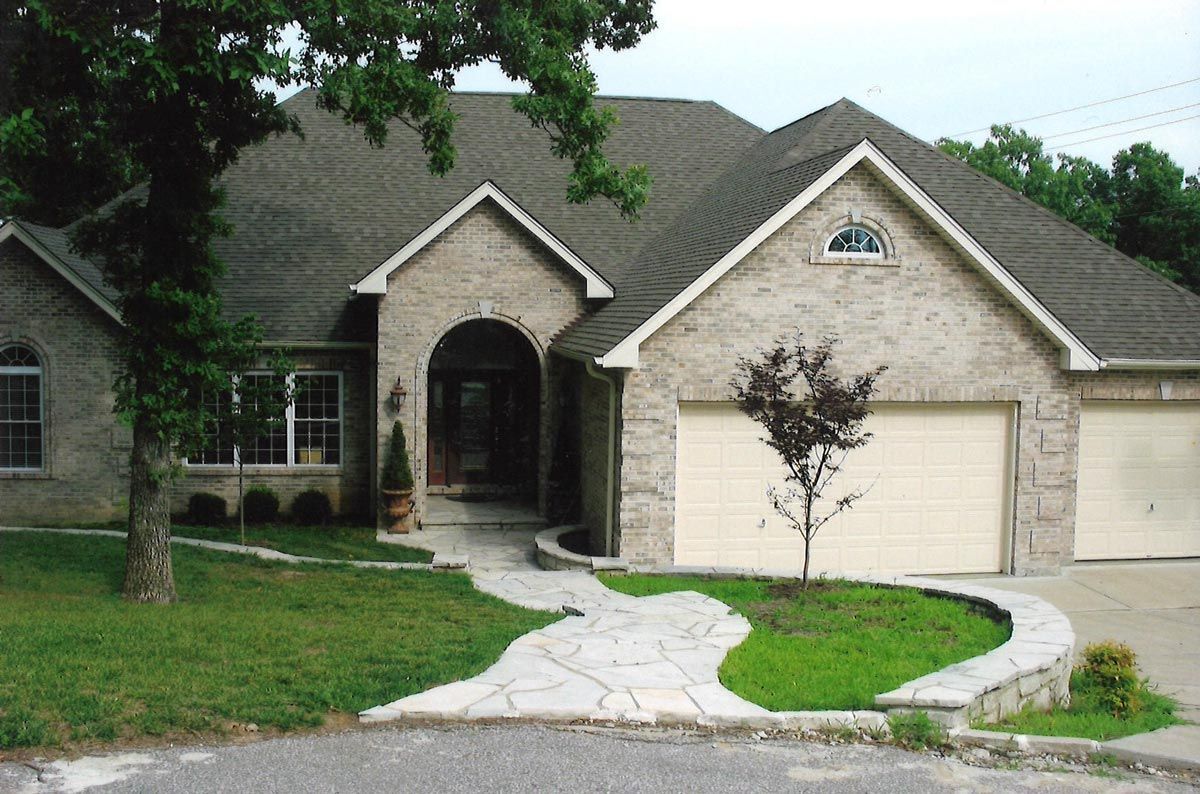 Gray brick house with a stone pathway leading to the arched front door, flanked by a tree and garage.