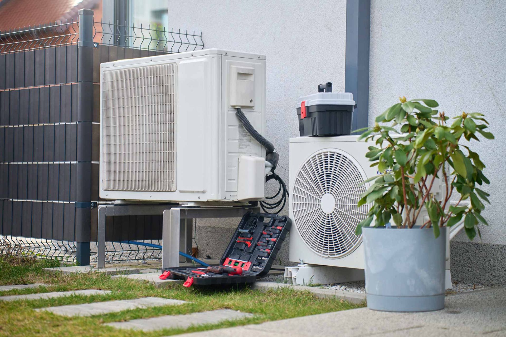 Outdoor AC units beside a fence, with a potted plant and tools on the grass near a house wall