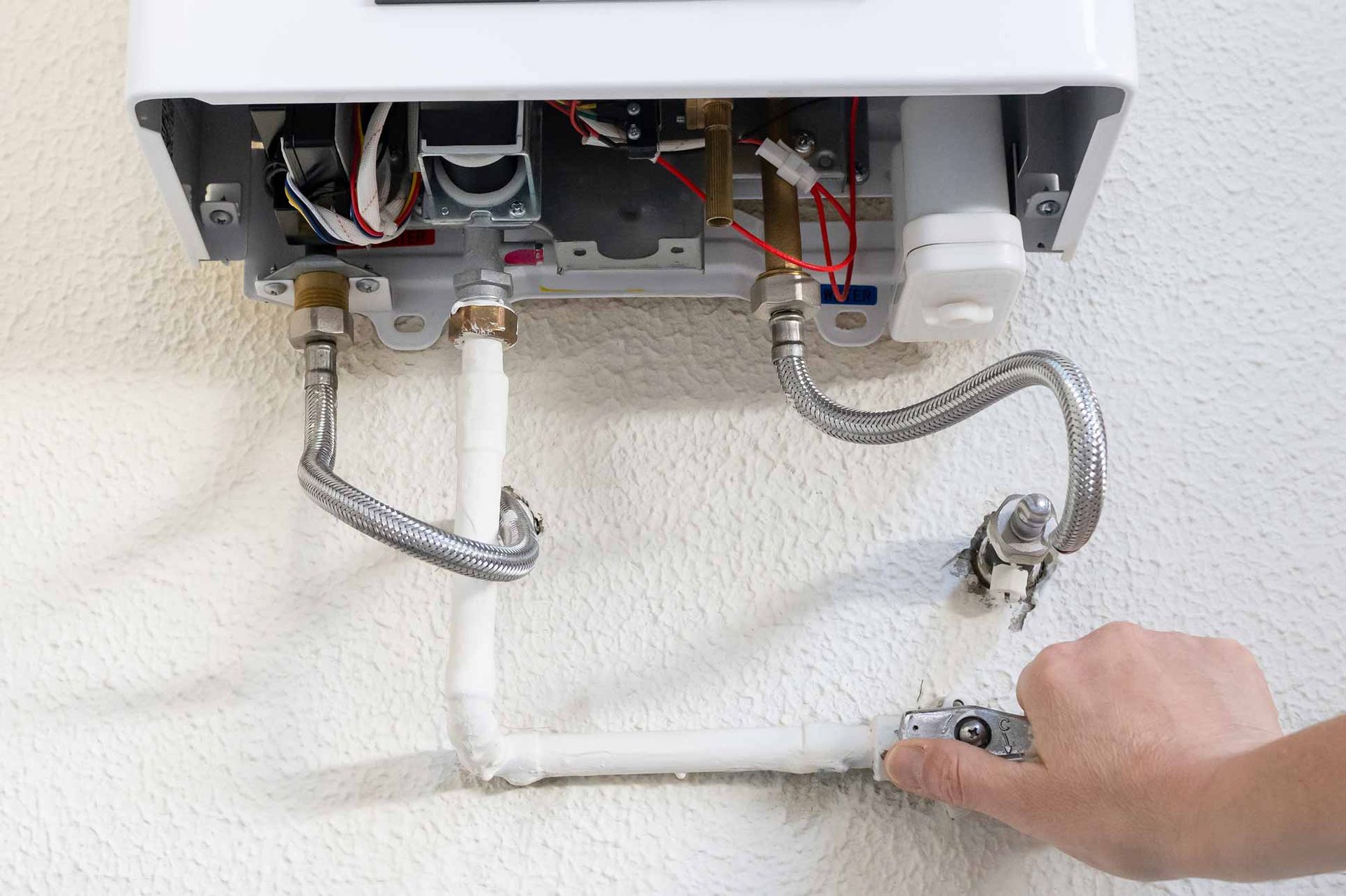 Technician tightening a pipe connection under a wall-mounted boiler, with exposed plumbing and wiring.