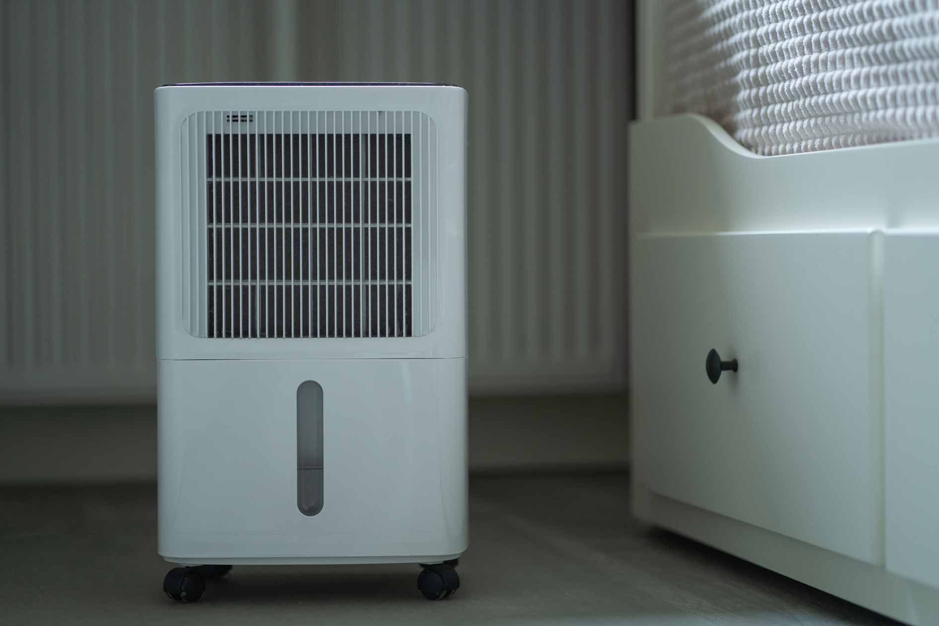 White portable dehumidifier on wheels beside a white bed in a bedroom