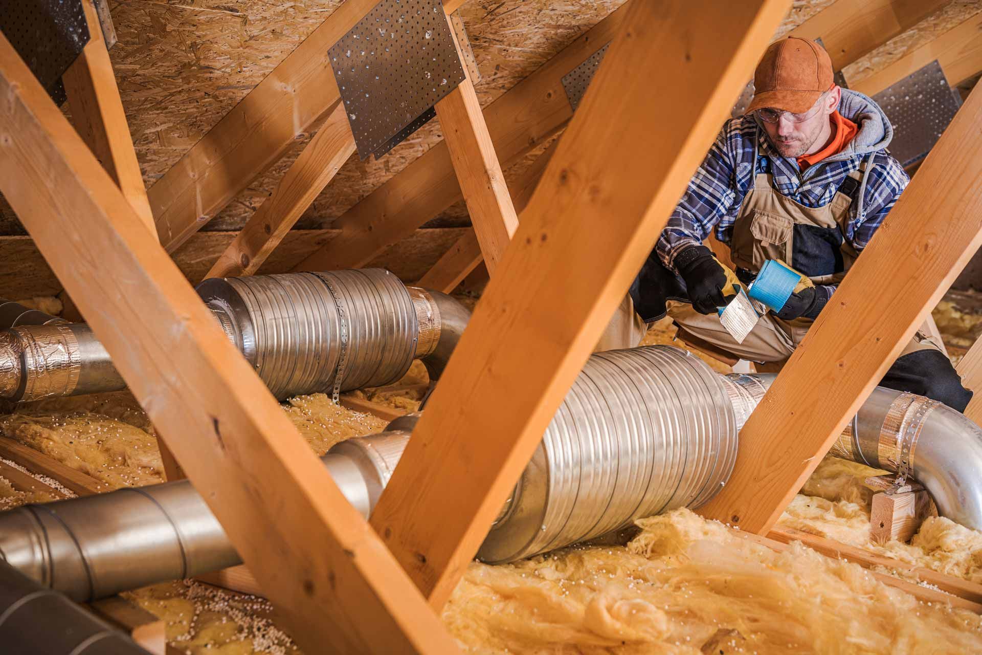 Worker in orange hard hat inspecting silver ducts in an attic with exposed wooden beams and insulation