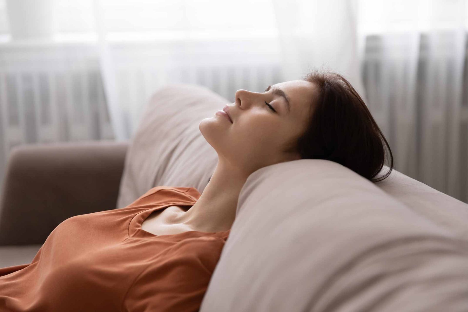 Person resting on a couch with eyes closed, wearing a peach shirt, in a bright room