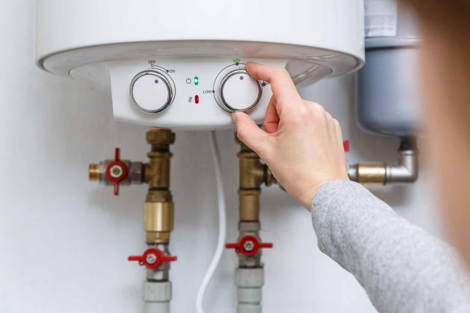 Hand adjusting a white water heater’s temperature knob above exposed pipes and valves