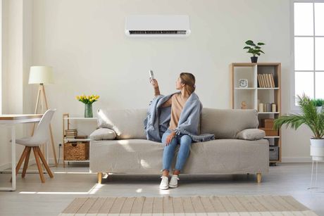 Person sitting on a gray sofa in a bright living room, holding a tablet near a wall-mounted air conditioner.