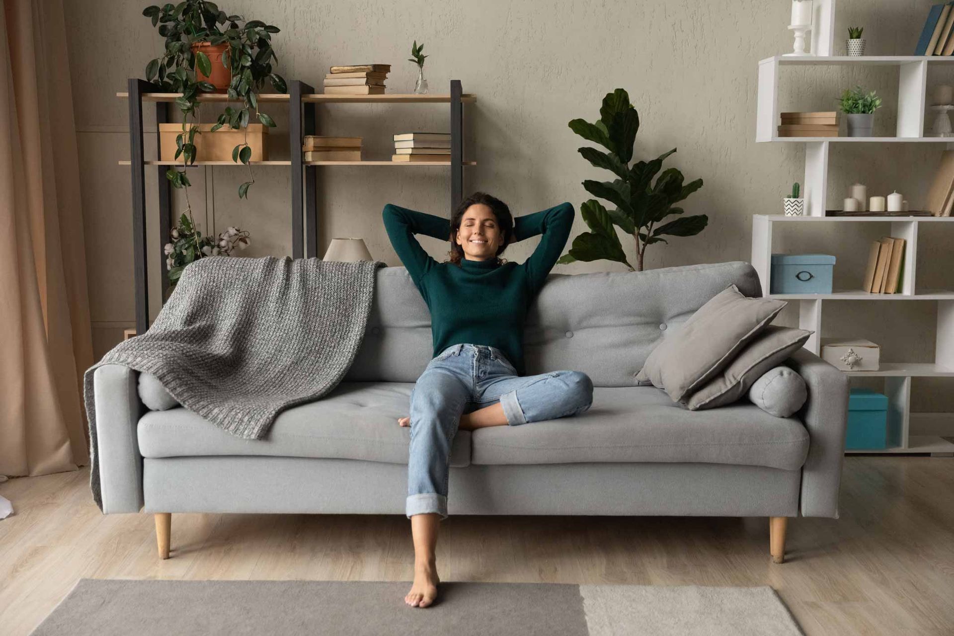 Person relaxing on a gray sofa in a cozy living room with plants and shelves.