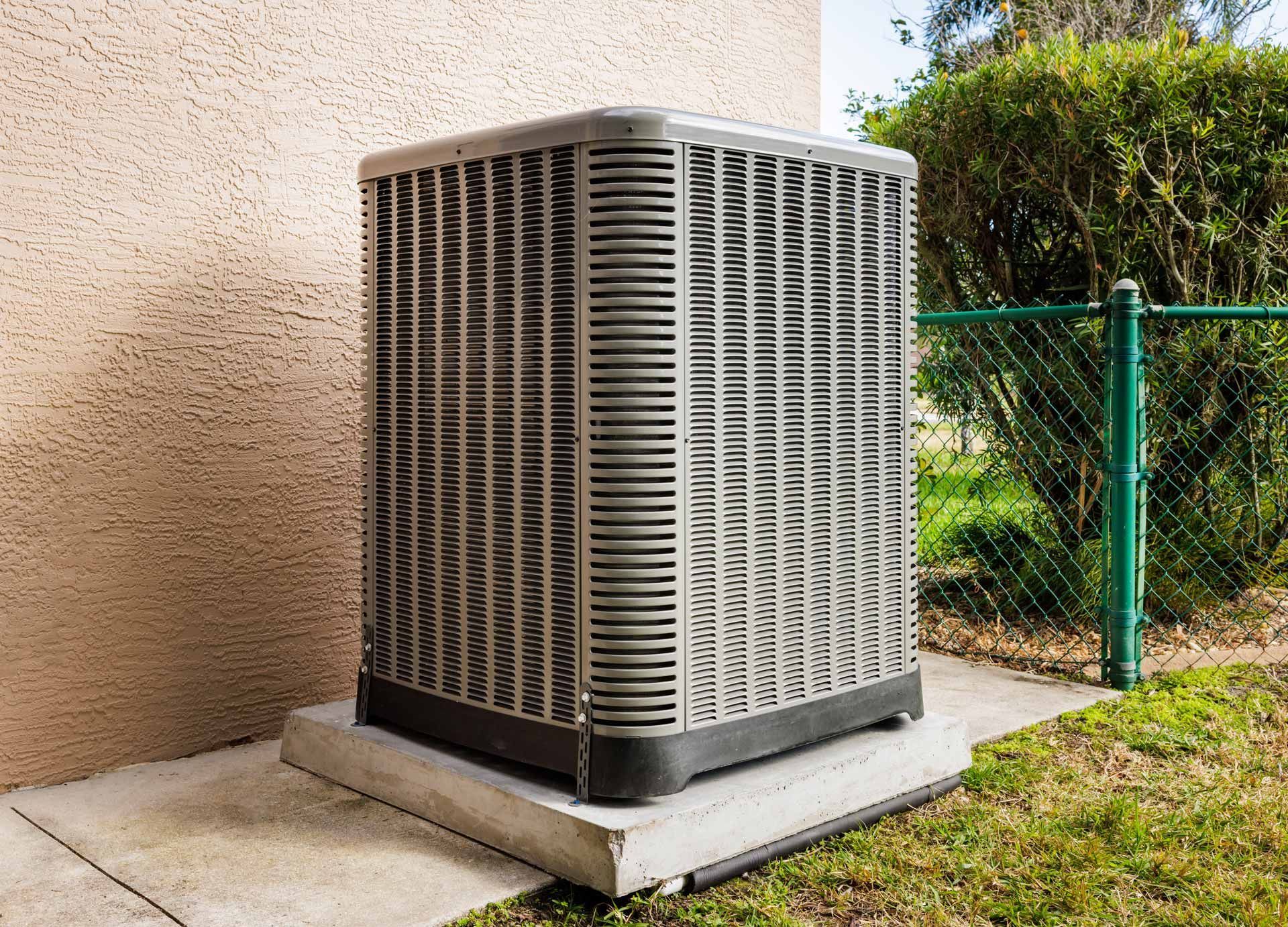 Outdoor air conditioner unit beside a stucco wall and green fence on a concrete pad