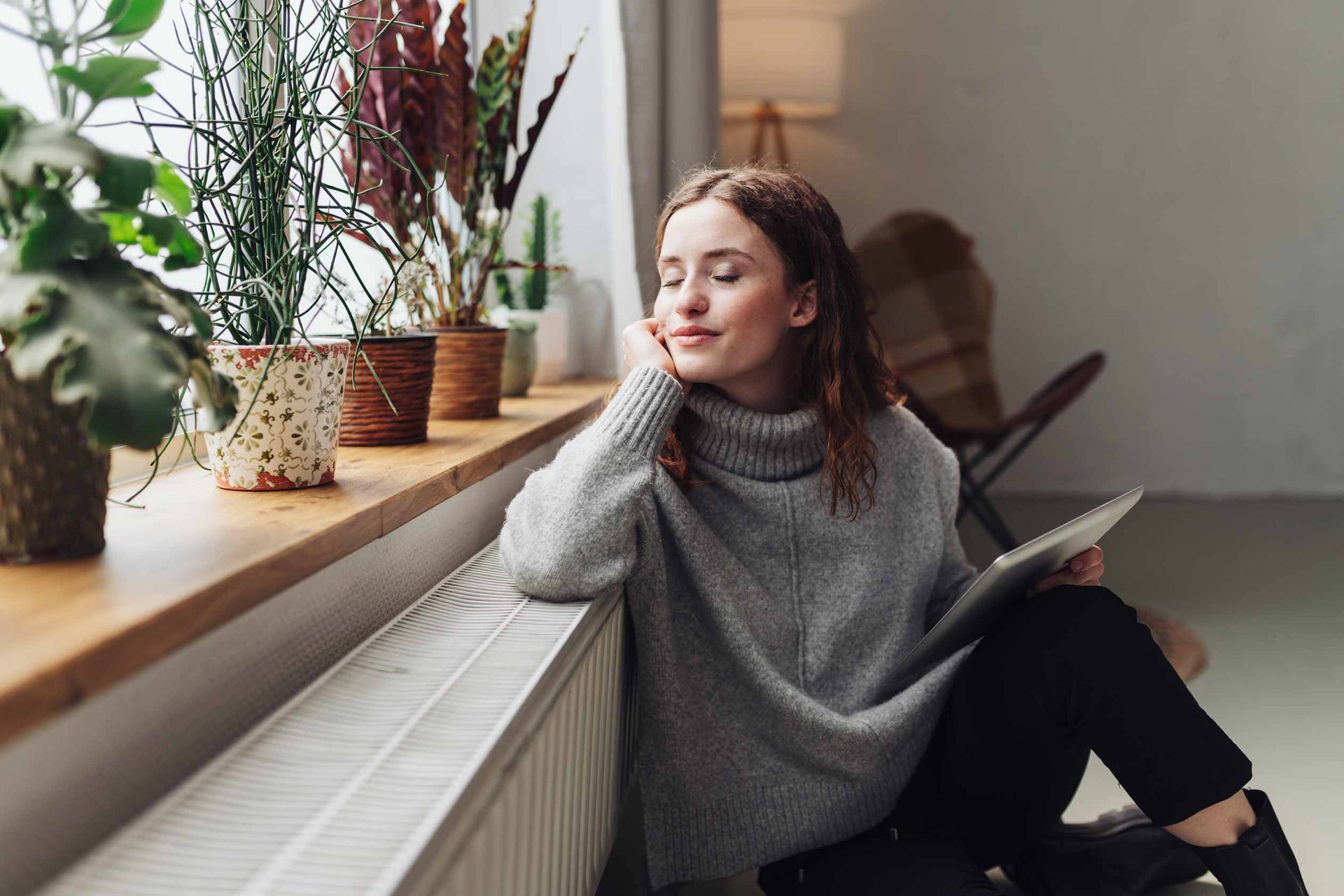 Person sitting by a window, resting their head on a hand beside potted plants and a book