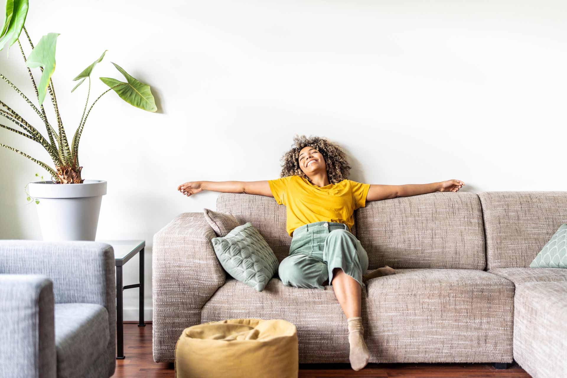 Person lounging on a beige sofa in a bright living room with a potted plant and yellow ottoman