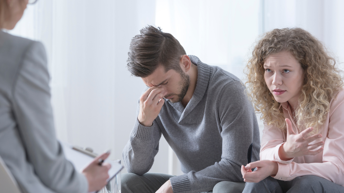 Couple in therapy, looking distressed, with a therapist in a neutral setting.