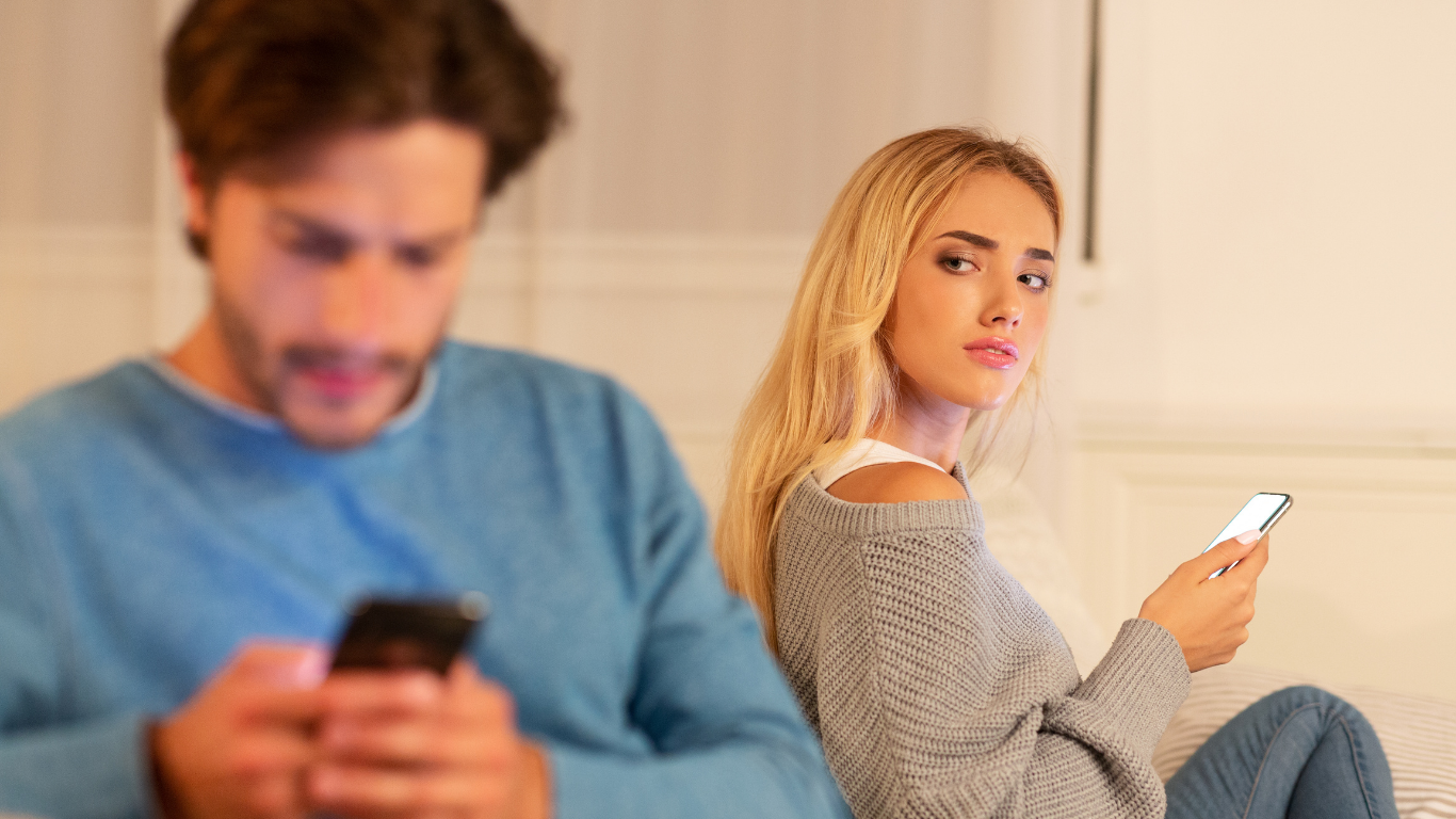 Woman looking at viewer while holding phone; man in background on his phone. Indoors.